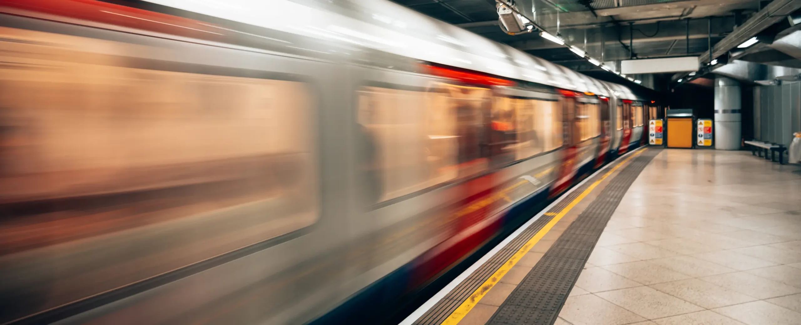 A motion-blurred train speeds through an underground station. The platform is empty, with bright lights reflecting off its clean, tiled floor, conveying haste.