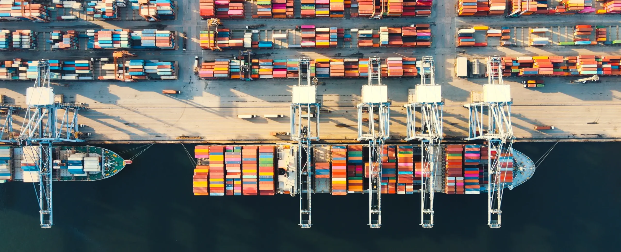 Aerial view of a busy port with colorful shipping containers in neat rows, large cranes, and two docked container ships, conveying industry and organization.