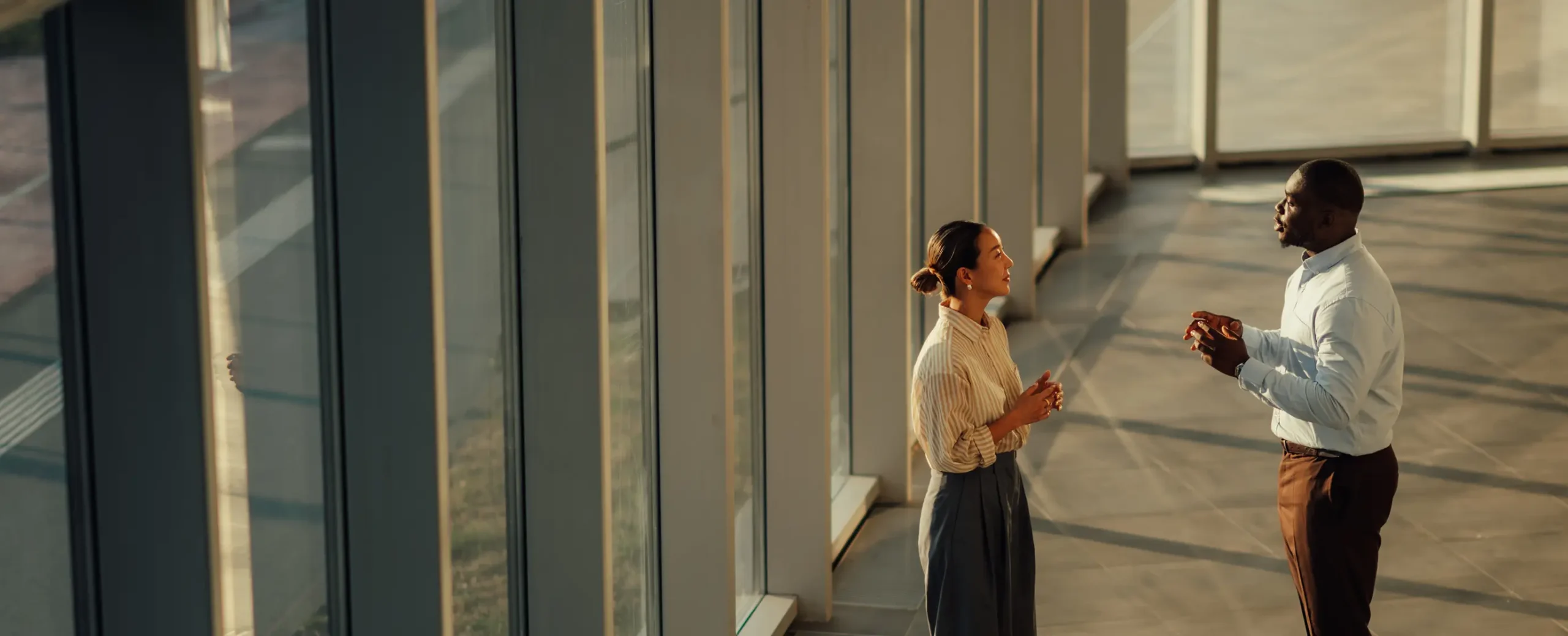 A man and woman converse in a sunlit, spacious room with large windows, casting long shadows. The setting conveys a professional and cordial atmosphere.