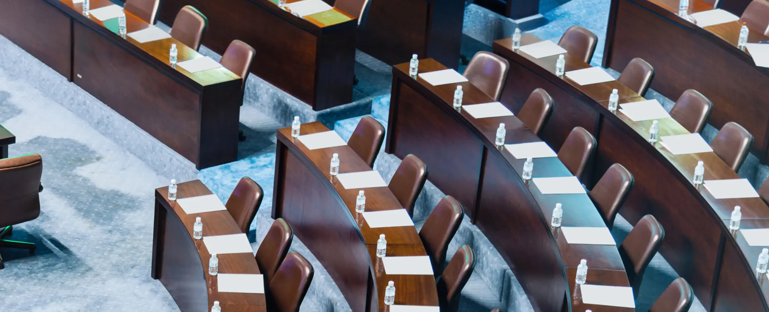 Rows of curved wooden desks in an empty auditorium, each with a notepad and water bottle, suggesting a formal setting or conference.