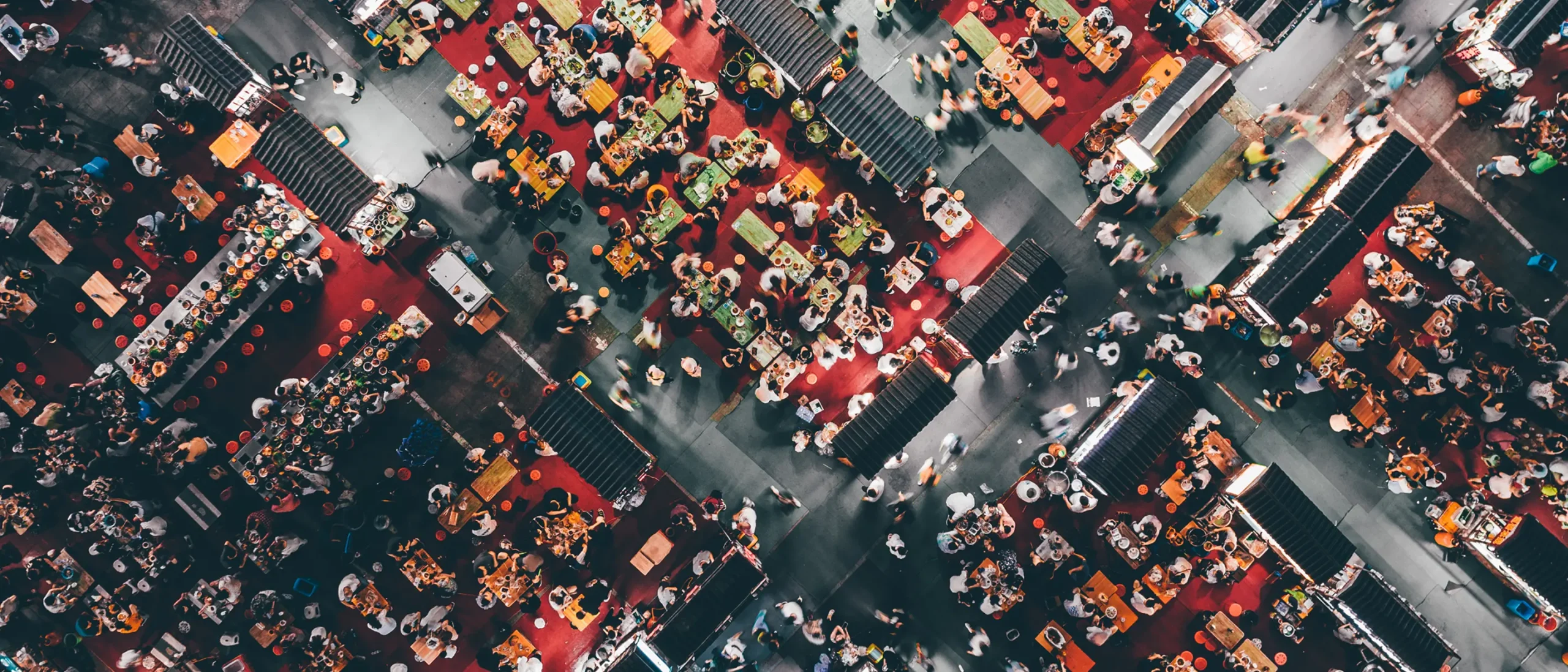 Aerial view of a bustling market with vibrant red and yellow stalls, numerous people shopping, and socializing. The scene conveys a lively, energetic atmosphere.