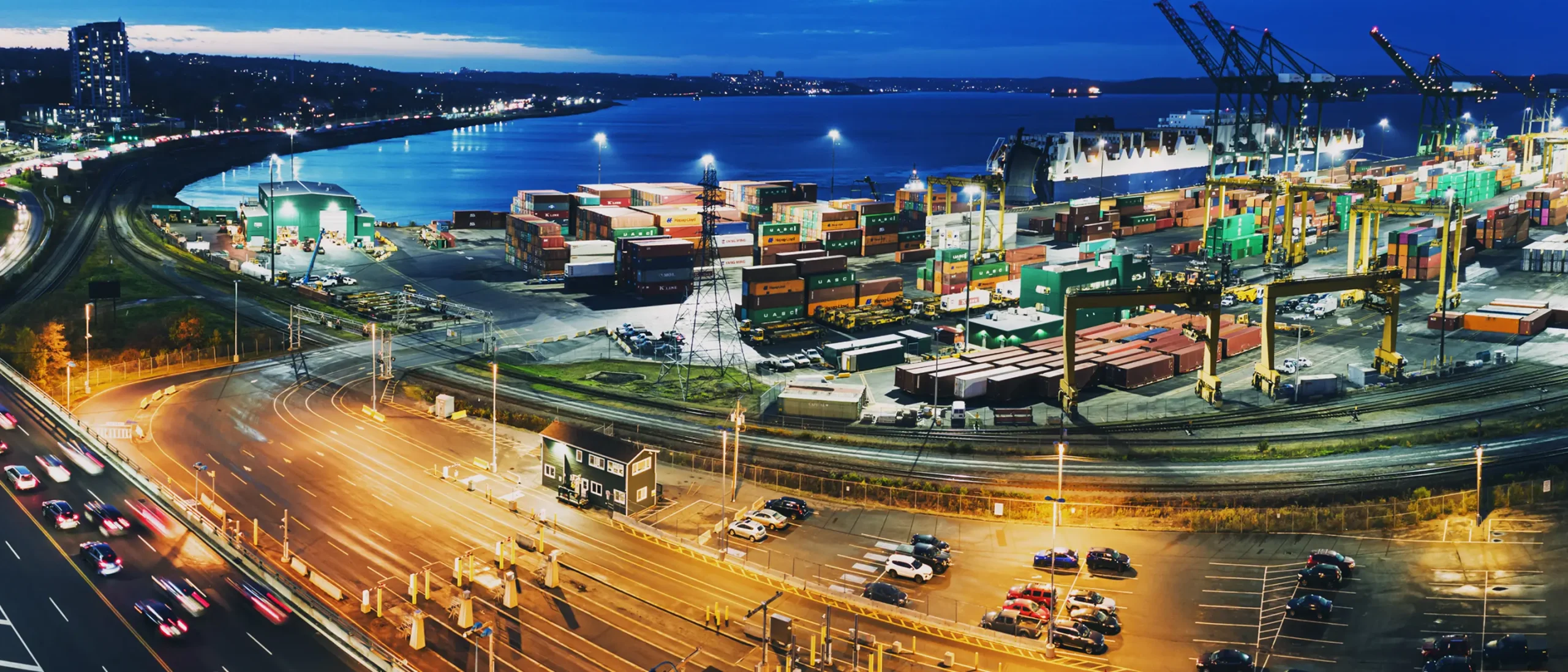 A vibrant coastal port at dusk, featuring colorful shipping containers, towering cranes, and bustling traffic on a nearby highway, illuminated under a deep blue sky.