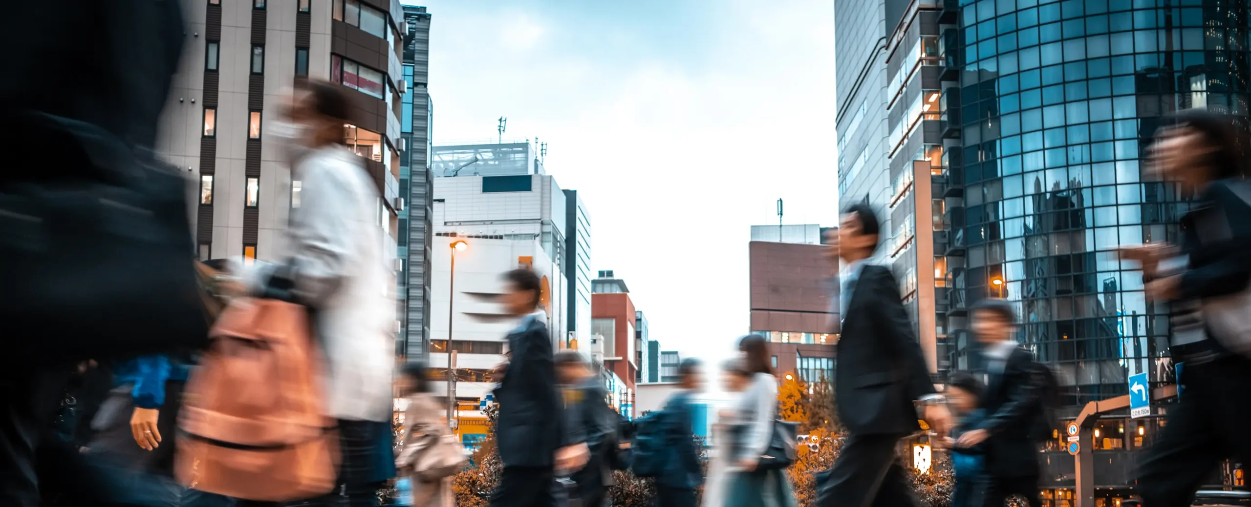 A bustling city street with blurred motion of people walking. Tall glass buildings surround, suggesting a busy, energetic urban environment.