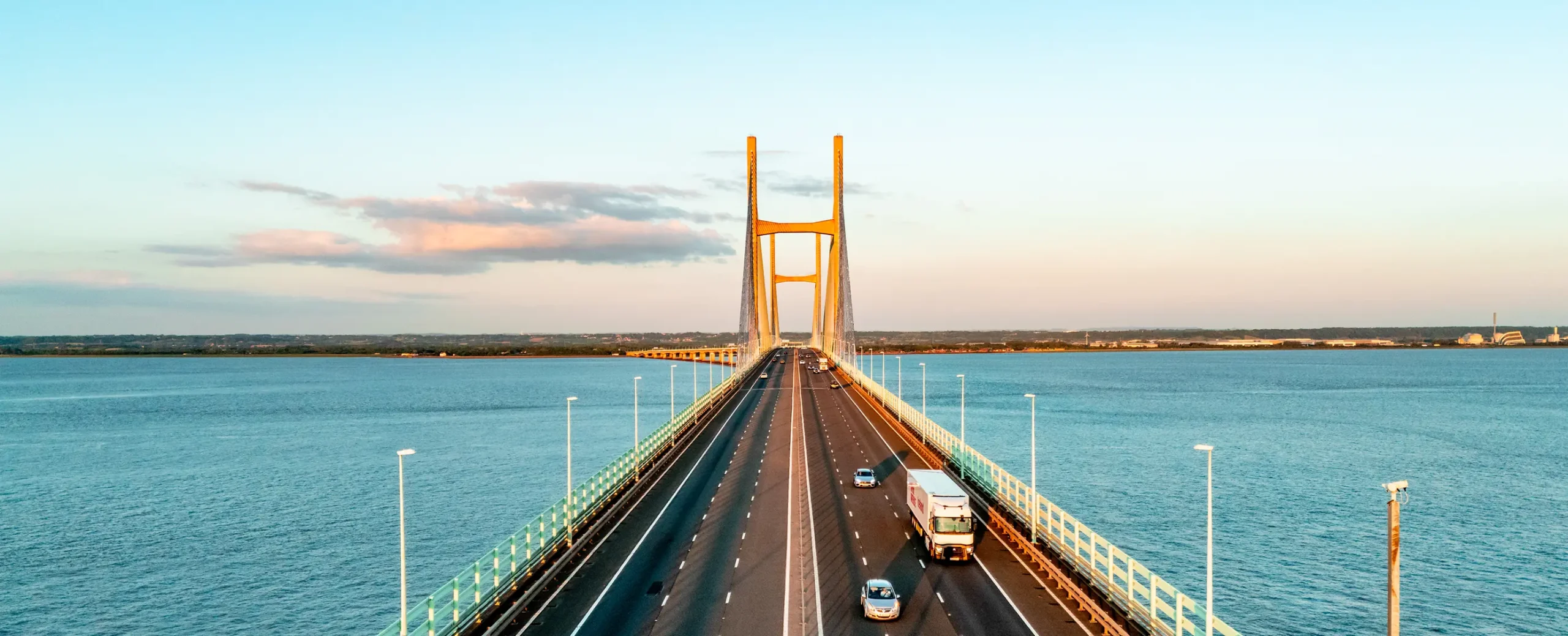 A wide suspension bridge stretches across calm blue water at sunset. Vehicles travel along the multi-lane road, conveying a sense of movement and tranquility.