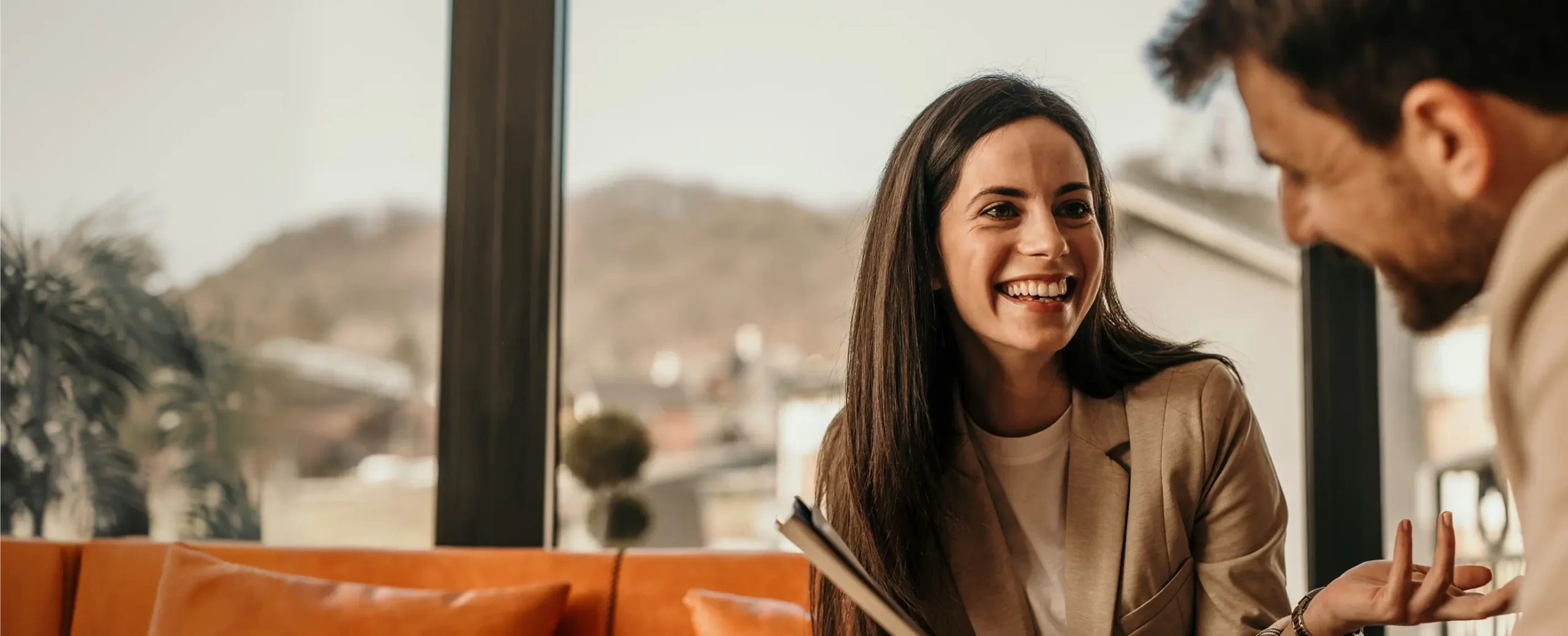A woman and a man are sitting on an orange sofa in a well-lit room, engaged in a friendly conversation. The woman is smiling, creating a warm, welcoming atmosphere.